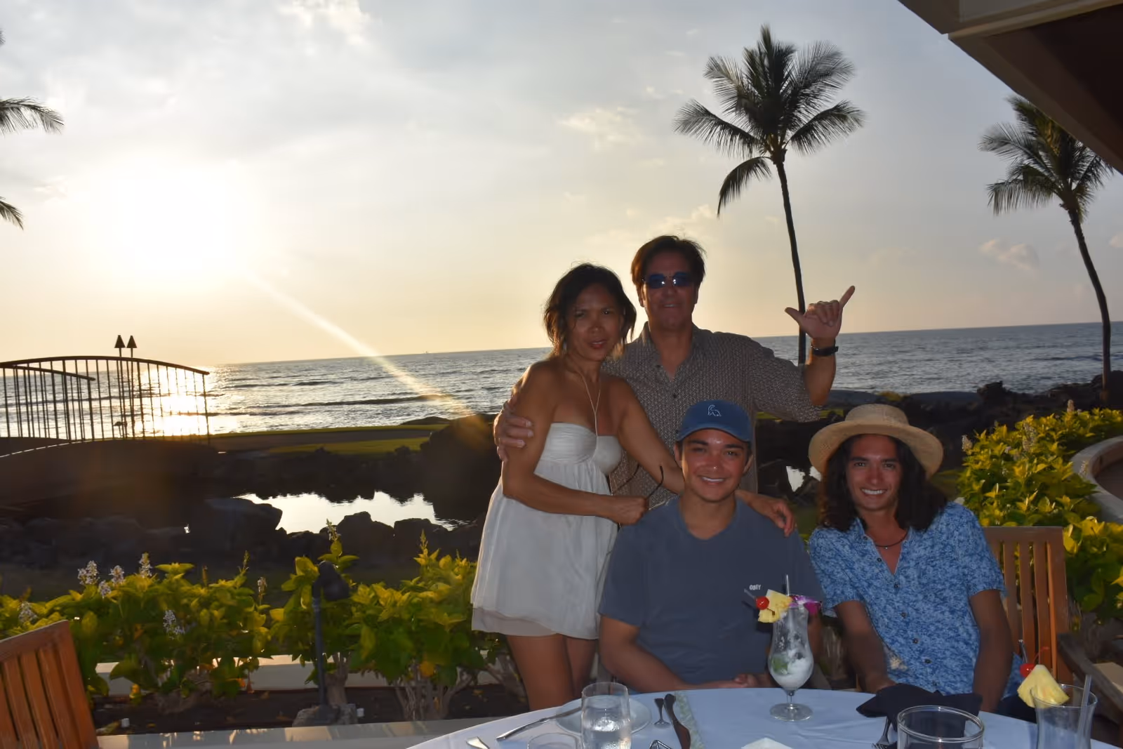 Four people posing and smiling at an outdoor dining table with tropical plants and palm trees in the background during sunset by the ocean. Two people are standing behind two seated individuals, one wearing a hat and the other a blue shirt. The setting appears relaxed and scenic.