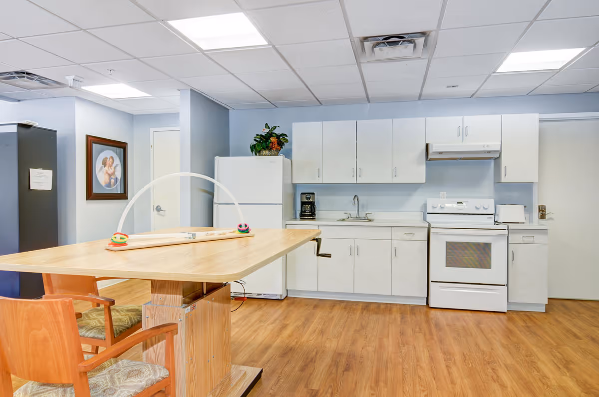 Bright communal kitchen with white cabinets, a refrigerator and stove, and a wooden table with chairs in the foreground.