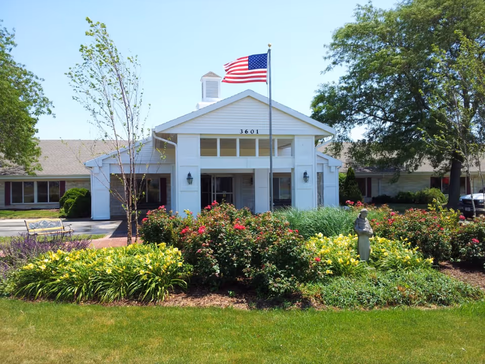 Front entrance of a single-story building with an American flag, the number 3601, flowering gardens and a small statue.