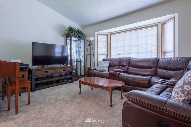Cozy living room with a brown leather sectional, wooden coffee table, TV on a stand, and a large bay window.