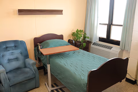 A small bedroom with a single bed covered with a green quilt, a blue upholstered armchair, a wooden overbed table, a potted plant on a nightstand, and a window with light blue curtains and an air conditioning unit below it.