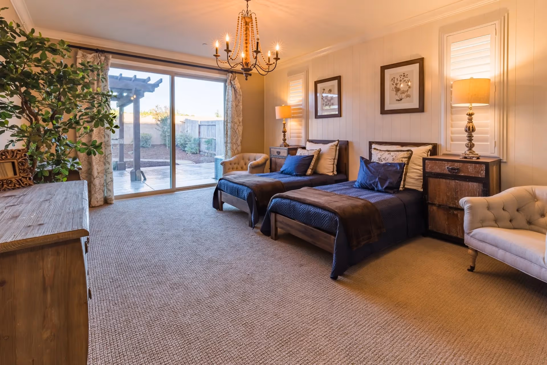 A cozy bedroom with two twin beds dressed in dark blue bedding and brown throws. Each bed has multiple pillows and is flanked by wooden nightstands with lamps. The room features a beige carpet, a chandelier hanging from the ceiling, a large sliding glass door leading to an outdoor patio, and a comfortable armchair. There are framed floral artworks on the wall and a large leafy plant near a wooden dresser.
