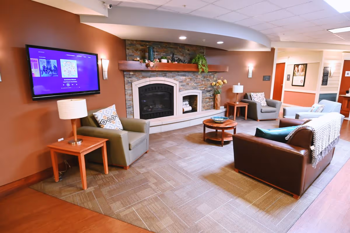 A cozy living room area with a stone fireplace, a mounted flat-screen TV on a brown wall, and several seating options including a green armchair, a gray armchair, a brown leather sofa with a throw blanket, and a light blue sofa. There are wooden side tables with lamps and a round wooden coffee table in the center. The room has a carpeted floor and soft lighting.