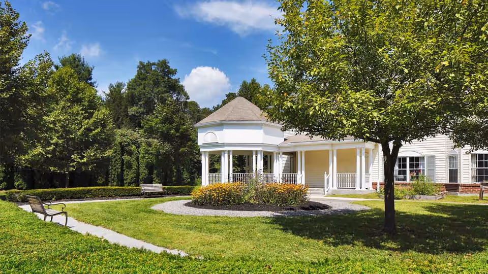 Outdoor garden area at The Cottage at Litchfield Hills featuring a white gazebo with a conical roof, surrounded by green grass, trees, flower beds, and benches under a blue sky with some clouds.