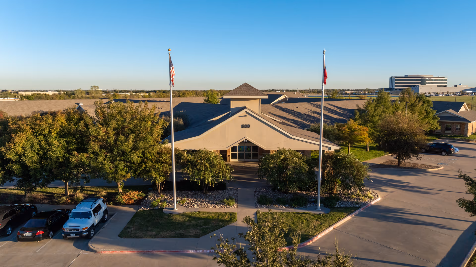 Front exterior view of Midlothian Healthcare Center building with two flagpoles displaying the American and Texas flags, surrounded by trees and parked cars in the parking lot under a clear blue sky.