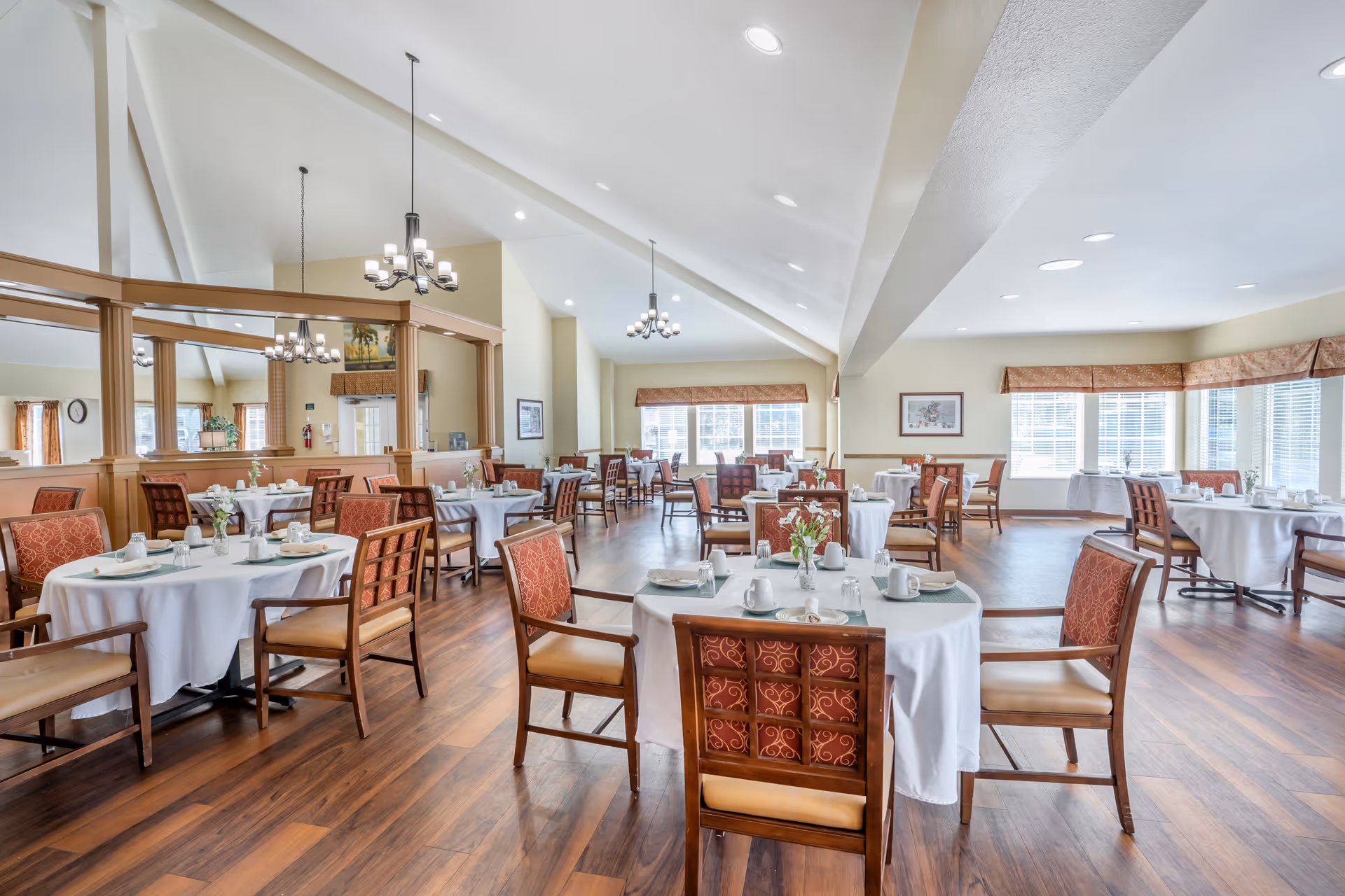Bright, spacious dining room with round tables set for meals, wooden chairs, chandeliers, and large windows.