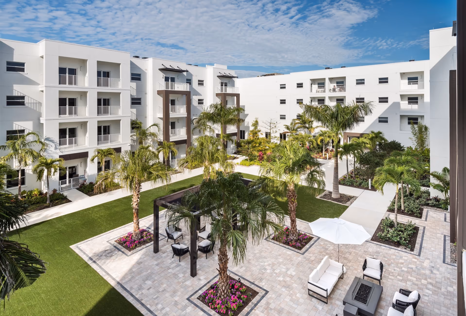 Outdoor courtyard area of a senior living facility with white multi-story buildings surrounding a landscaped garden. The courtyard features palm trees, green lawns, paved walkways, a seating area with white cushioned chairs and sofas, a white umbrella, and a pergola structure.