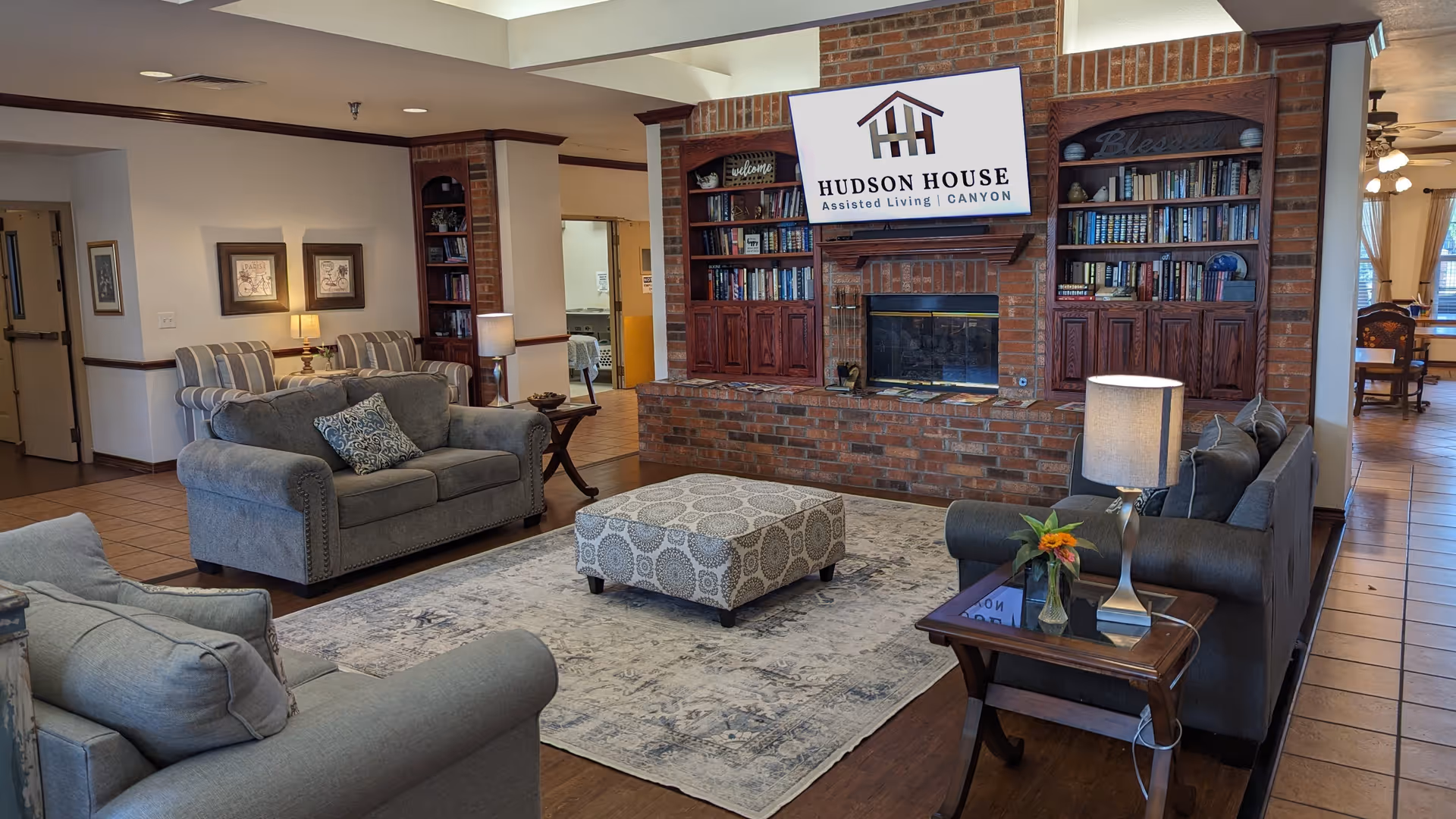 A cozy living room area in Hudson House Canyon assisted living facility featuring three gray upholstered sofas arranged around a patterned ottoman on a large area rug. The room has a brick fireplace with built-in wooden bookshelves on either side filled with books and decorative items. A sign above the fireplace reads 'Hudson House Assisted Living Canyon.' There are table lamps on side tables next to the sofas, and the room has warm lighting with a tiled floor and an open doorway leading to another room.