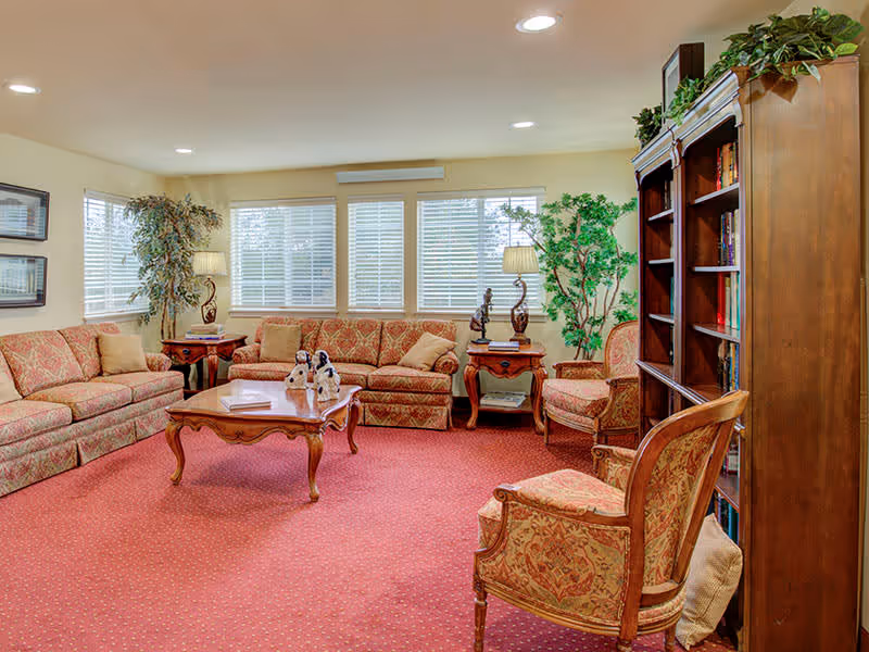 A cozy living room with patterned upholstered sofas and armchairs arranged around a wooden coffee table. The room has large windows with white blinds, two side tables with lamps and decorative sculptures, a tall wooden bookshelf filled with books, and green potted plants in the corners. The carpet is red with a subtle pattern, and the walls are painted a light beige color.