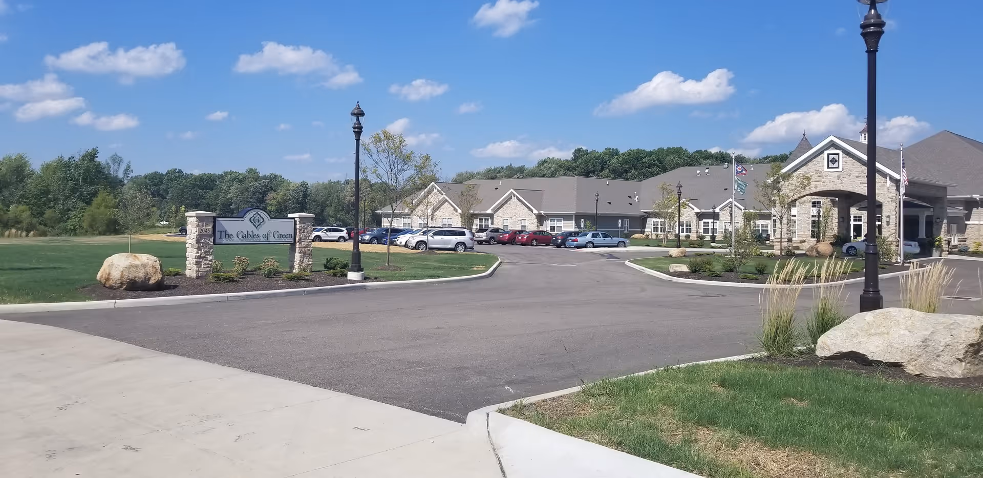 Exterior view of The Gables of Green senior living facility showing a large parking area, landscaped grounds with grass and rocks, a stone sign with the facility name, and a building with a covered entrance under a blue sky with scattered clouds.