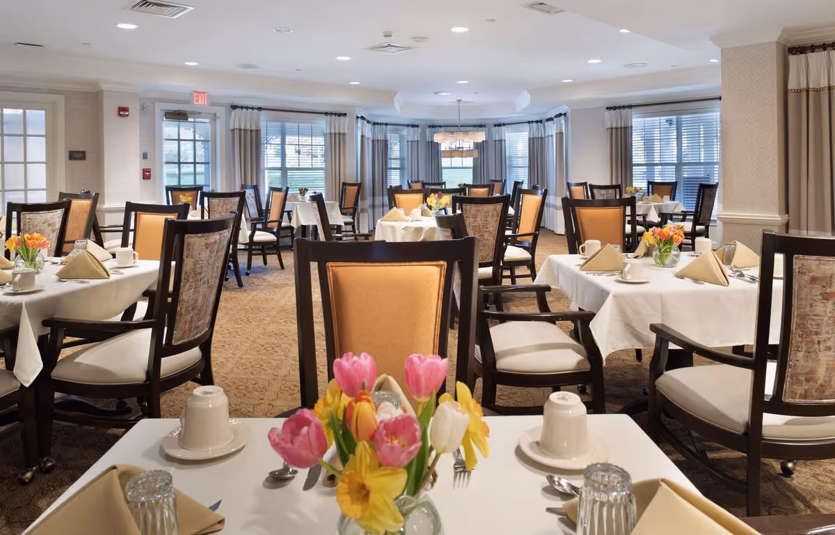 Well-lit dining room with multiple tables set with white tablecloths, floral centerpieces, and upholstered chairs.