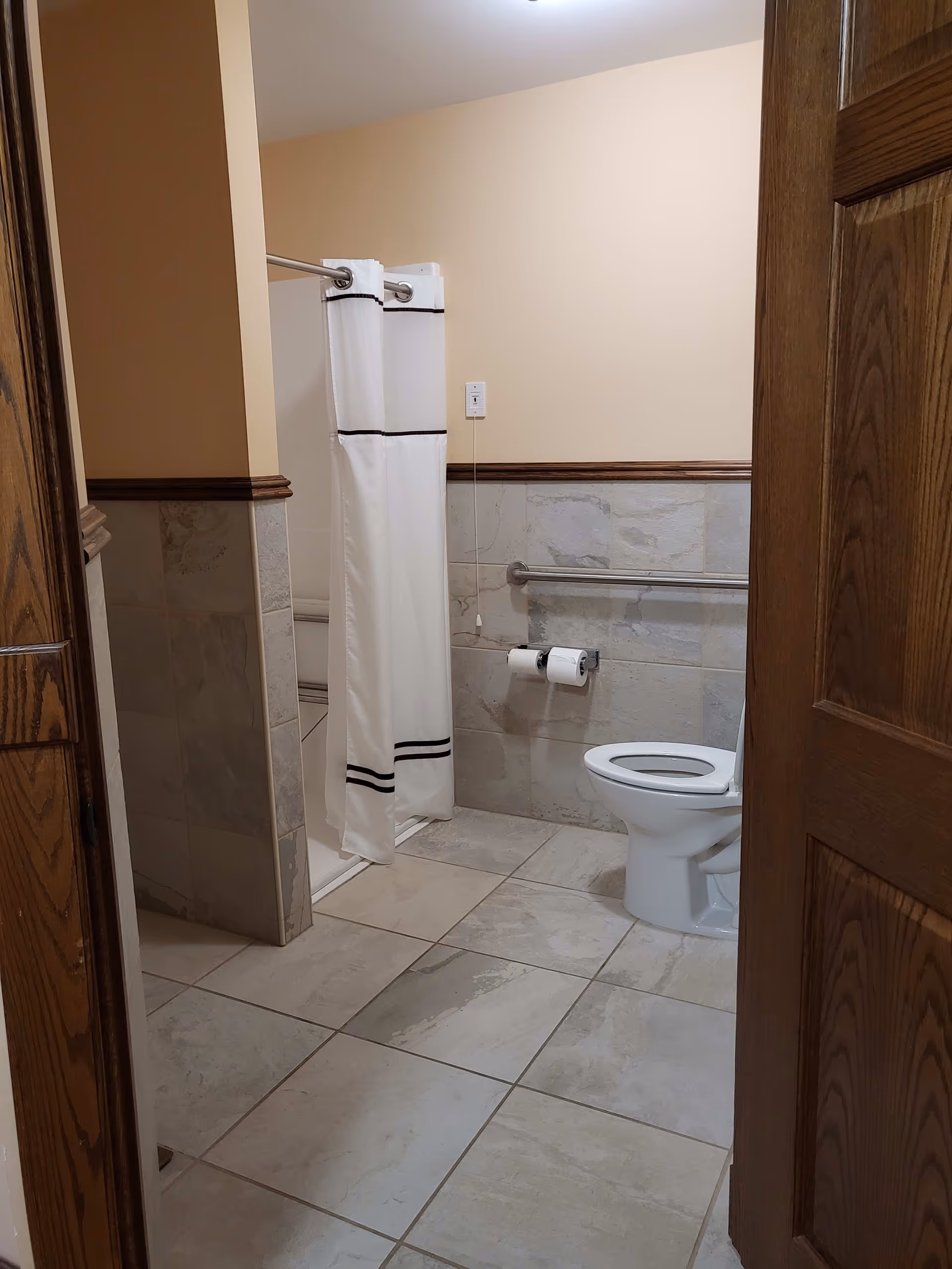 A bathroom with beige walls and tiled floor and lower walls. There is a white toilet with a grab bar on the wall behind it, two rolls of toilet paper on a holder, and a shower area with a white curtain featuring two black horizontal stripes near the top and bottom. The door to the bathroom is wooden and partially open.