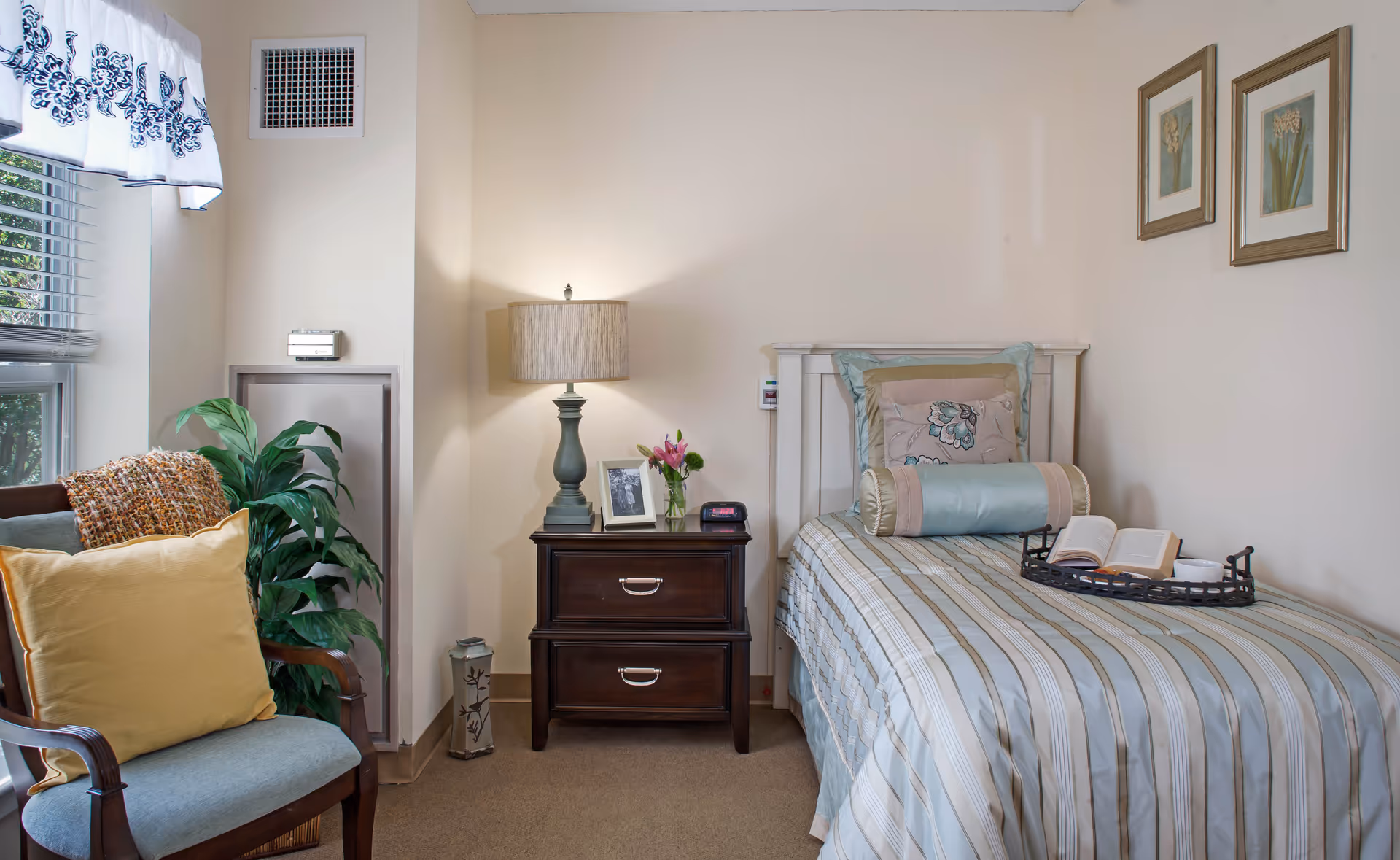 A cozy bedroom in a senior living facility featuring a single bed with striped bedding and decorative pillows. A dark wooden nightstand beside the bed holds a lamp, a framed photo, a small vase with flowers, and a clock. A wooden chair with a yellow cushion and a knitted throw is placed near a window with white and blue patterned curtains. Two framed floral artworks hang on the wall above the bed.