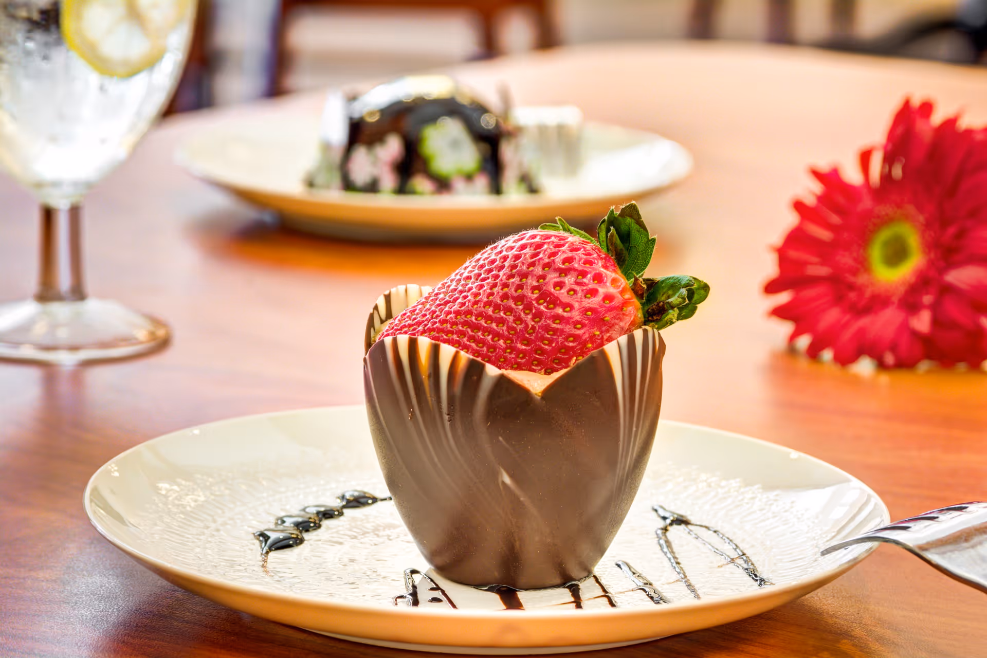 A large strawberry served inside a chocolate cup on a decorated dessert plate on a wooden table with a drink and flower in the background.