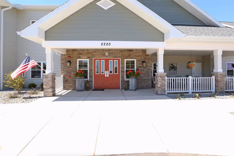 Front entrance of a senior living facility with a covered porch, stone pillars, and a red double door. The building number 2220 is displayed above the door. There are two large planters with red flowers on either side of the entrance and an American flag on the left side. The exterior walls are light gray with white trim.