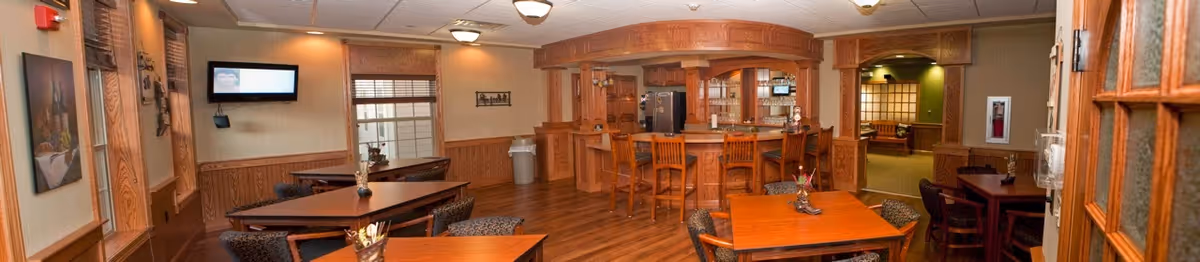 Interior view of a common area in a retirement community featuring wooden tables and chairs, a bar counter with high stools, wood-paneled walls, and warm lighting. There are decorative items on the tables and framed artwork on the walls, with a television mounted on one wall and a hallway leading to another room.