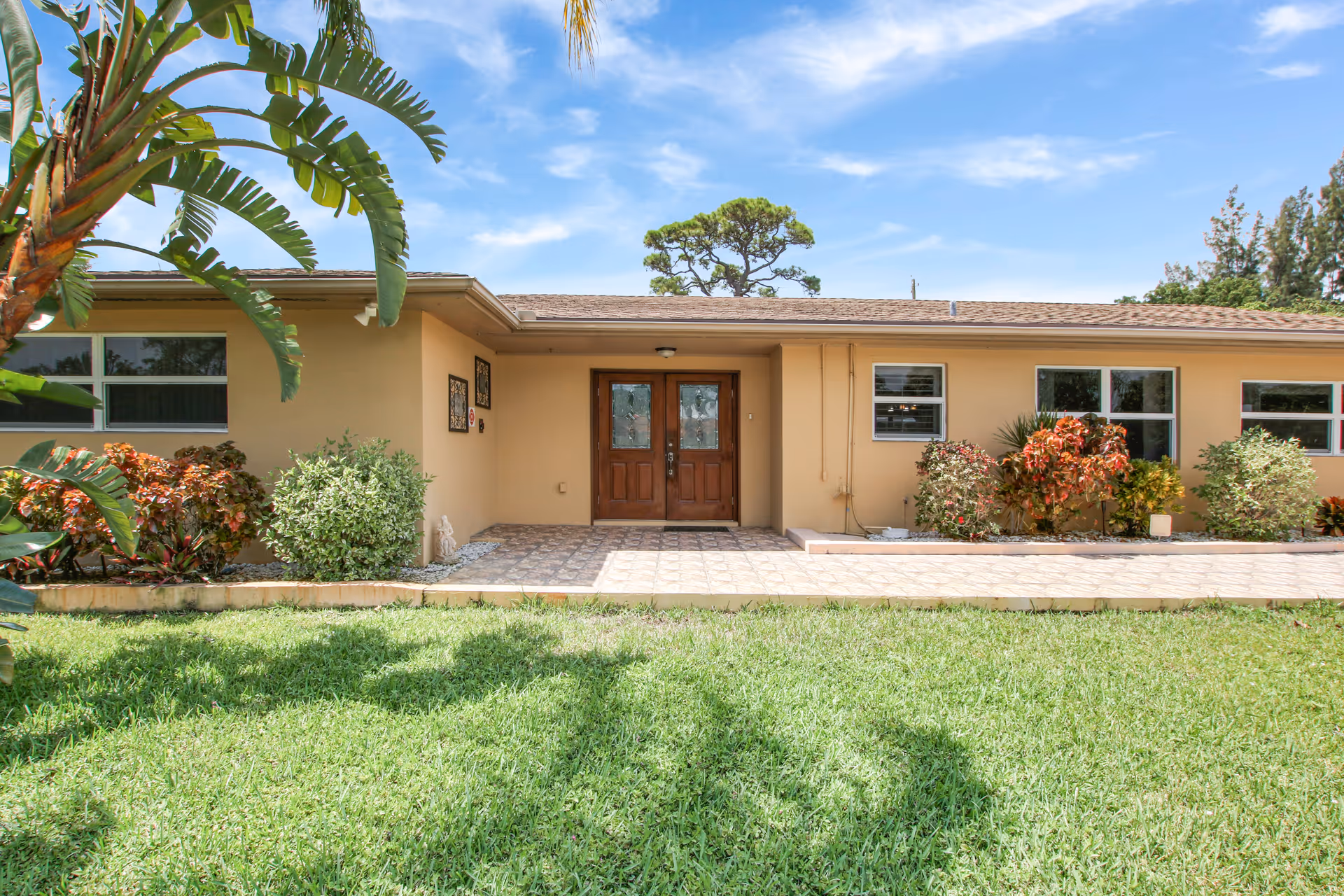 Front exterior view of Hibiscus Palace Assisted Living Facility showing a single-story building with a tiled entrance, wooden double doors, several windows, and landscaped greenery including bushes and palm trees under a blue sky with some clouds.