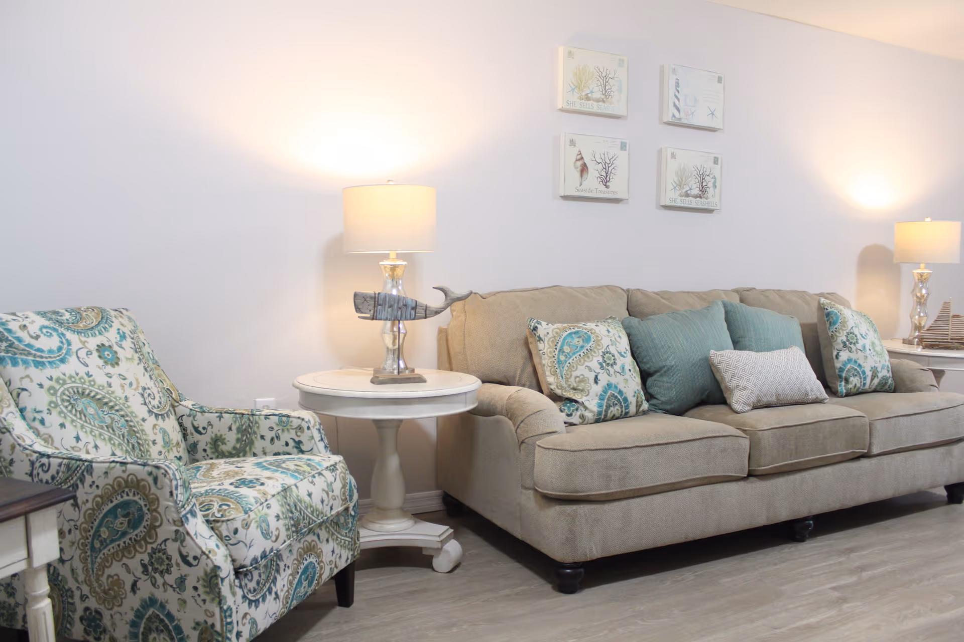 A cozy living room with a beige sofa adorned with patterned and solid teal cushions, a patterned armchair, two white side tables each with a lamp, and four framed sea-themed artworks on the wall.