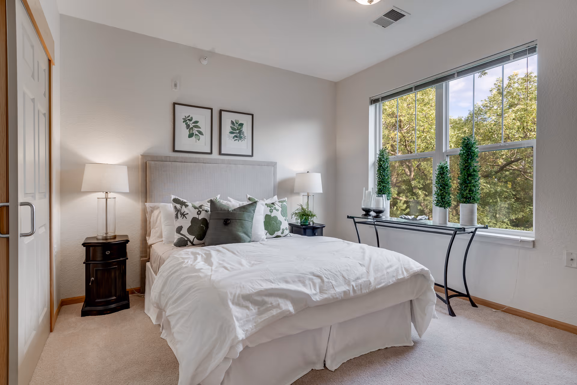 Bright furnished bedroom with a bed, bedside tables and lamps, botanical artwork and a large window with potted plants.