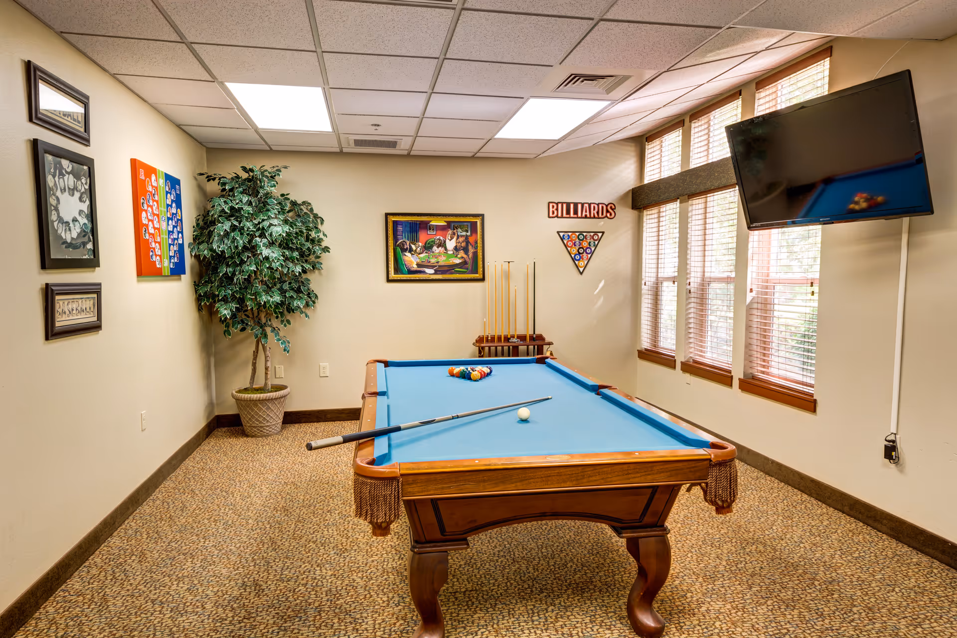 A well-lit game room featuring a billiards table with a blue felt surface, a cue stick resting on the table, and a rack of billiard balls arranged for a game. The room has beige walls adorned with framed pictures and a 'BILLIARDS' sign above a rack holding pool cues. There is a large potted plant in the corner and a flat-screen TV mounted on the wall next to windows with wooden blinds.