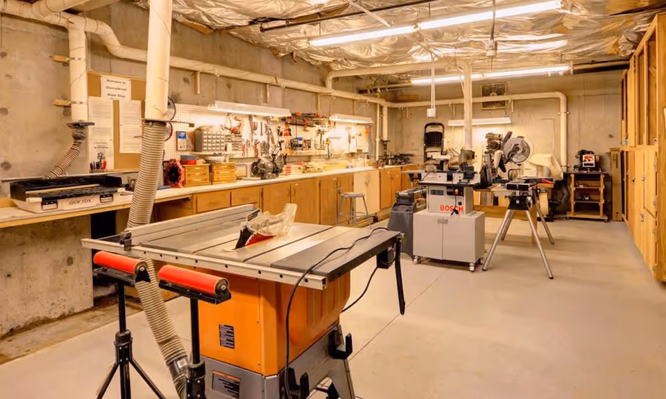 A well-organized workshop room with various woodworking tools and equipment including a table saw, miter saw, and other power tools. The room has wooden cabinets along the walls, a concrete floor, and exposed ceiling pipes covered with insulation. There are workbenches with tools and supplies neatly arranged.