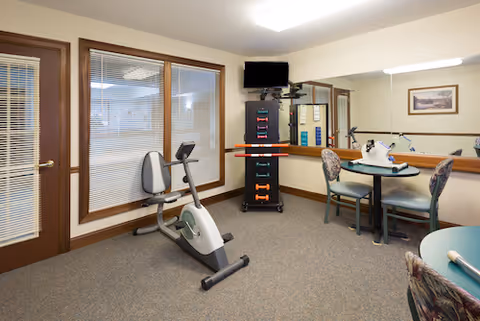 A small exercise room with a stationary bike, a rack of dumbbells, a wall-mounted TV, and a table with four chairs. The room has beige walls, carpeted floor, and large windows with blinds. A framed picture hangs on the wall above the table.