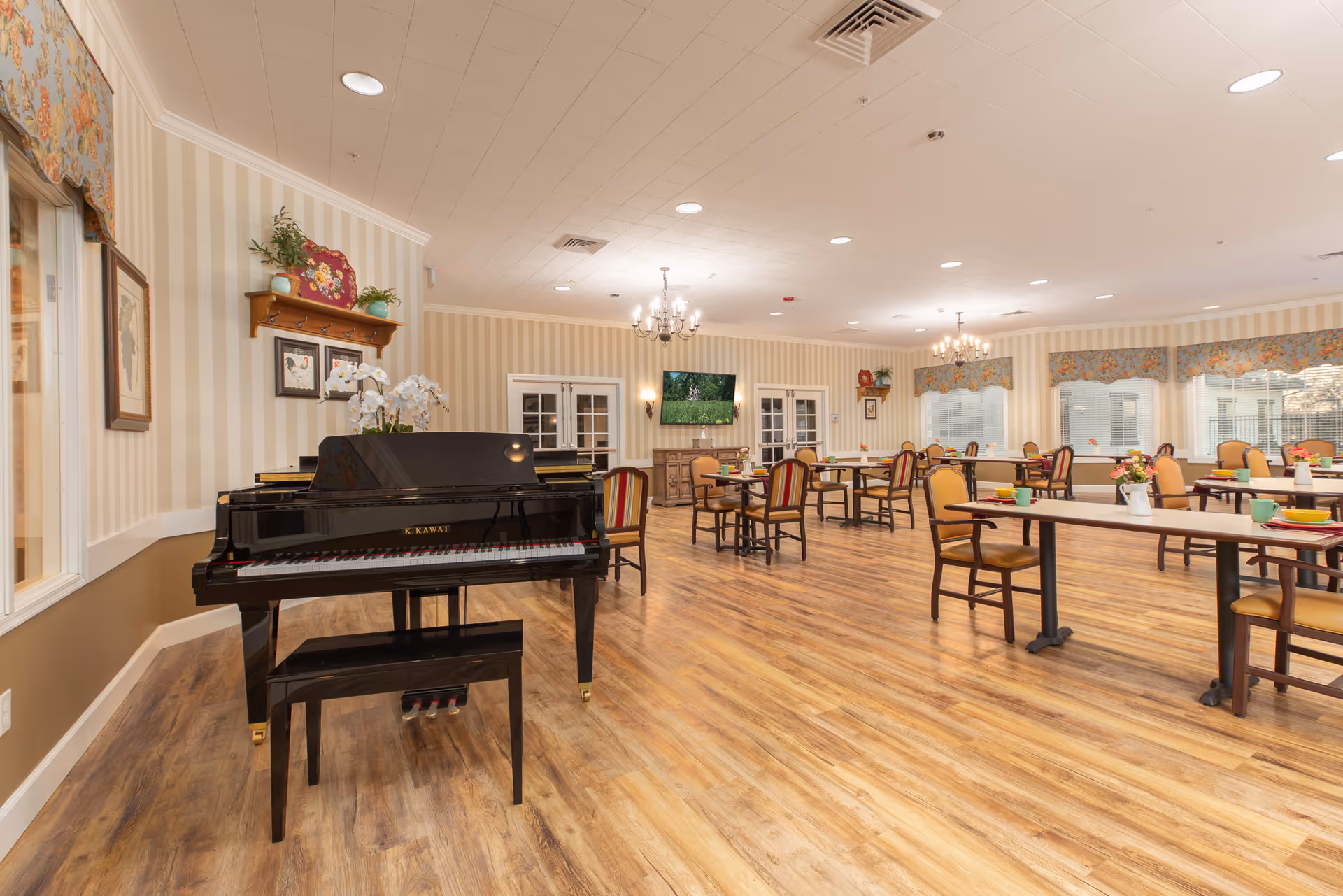 A spacious dining room with wooden floors, multiple tables and chairs arranged neatly, a black grand piano with a bench in the foreground, floral curtains on the windows, chandeliers hanging from the ceiling, and a wall-mounted TV in the background.