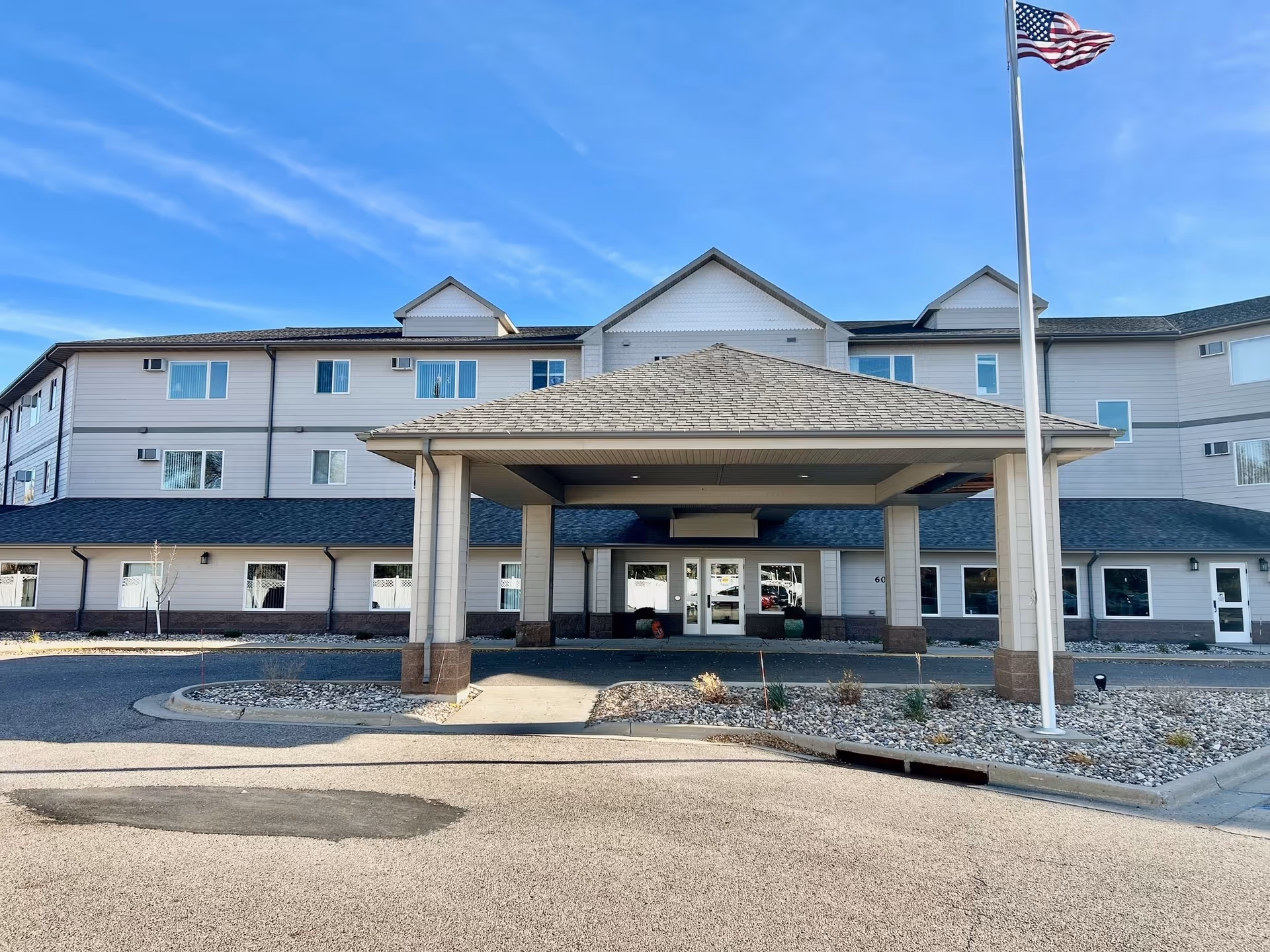 Exterior front view of Minot Lodge Senior Living building with a covered entrance, multiple windows, and an American flag on a flagpole in front. The sky is clear and blue.