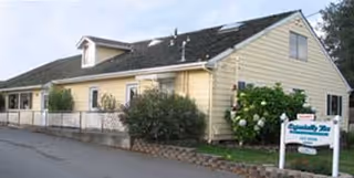 Exterior view of a single-story yellow building with a dark roof, surrounded by bushes and trees. There is a white sign in front of the building that reads 'Especially You Assisted Living'.