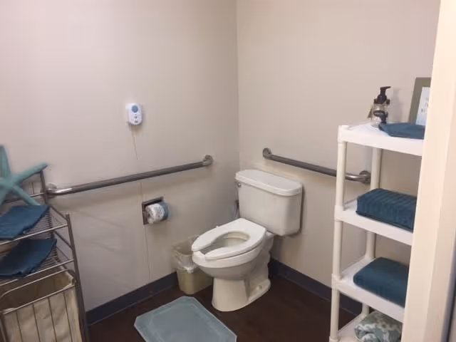 A small bathroom with a white toilet, grab bars on the walls, a metal rack with folded blue towels and a starfish decoration, a white shelving unit with folded blue towels and a soap dispenser, a small trash can, and a blue bath mat on the floor.