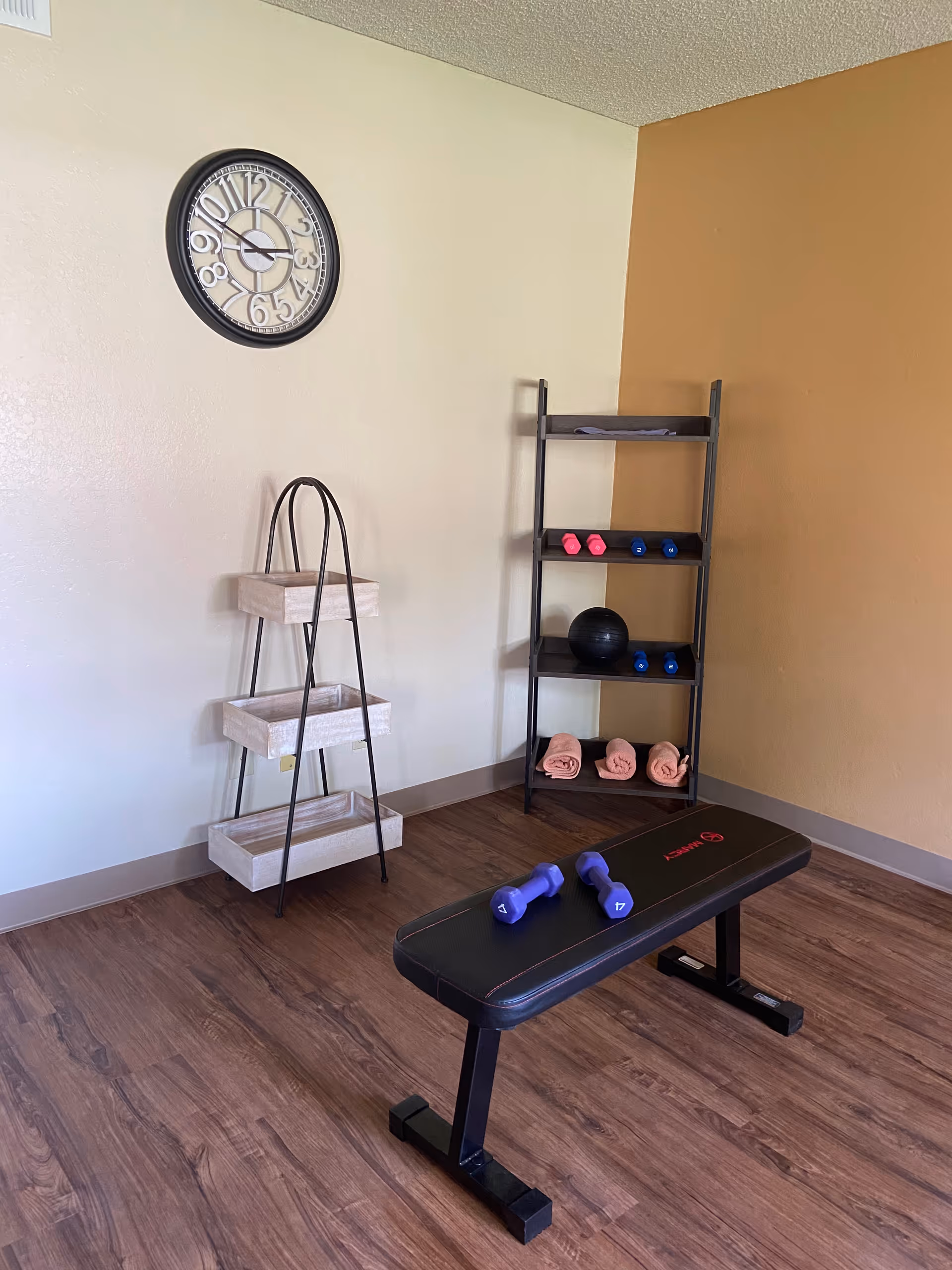 A small exercise room with a black workout bench holding two purple dumbbells. In the corner, there is a black metal shelf with pink towels, a black exercise ball, and several pairs of small dumbbells in pink and blue. A three-tier wooden and metal storage rack is against the wall. A round wall clock with large numbers hangs on the light-colored wall above the storage rack. The floor is wood laminate, and one wall is painted a warm tan color.