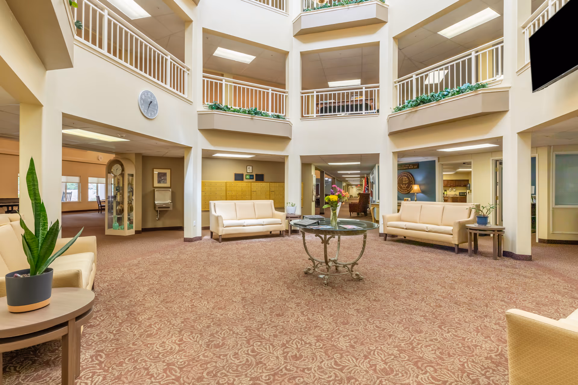 Spacious senior living community common area with beige sofas arranged around a central glass table holding a vase of flowers. The room has a high ceiling with a second-floor balcony railing, patterned carpet, and several potted plants. There is a clock on the wall, a water fountain, and a hallway leading further into the building.
