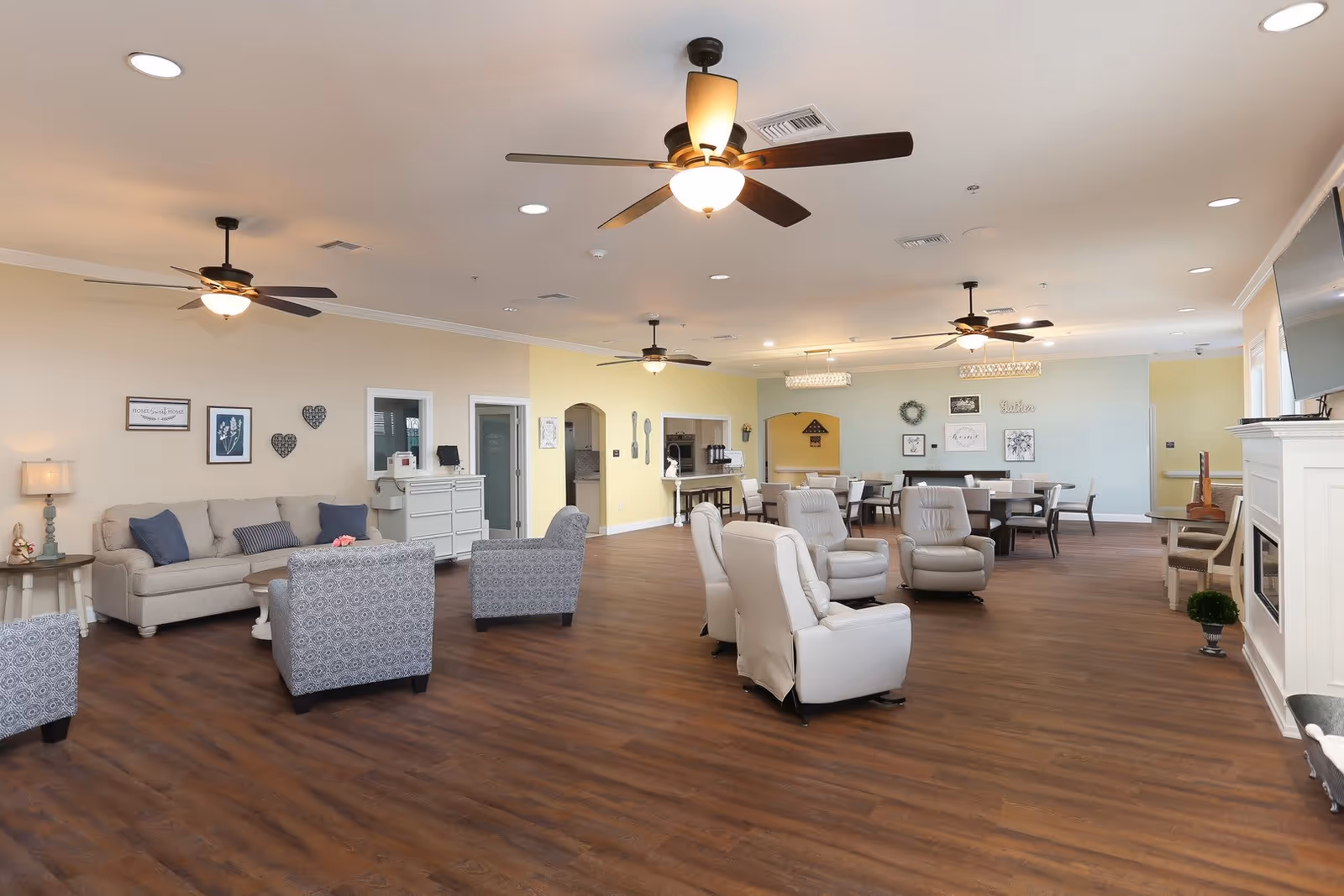 A spacious and well-lit assisted living common area featuring multiple seating options including a beige sofa with blue cushions, patterned armchairs, and white recliners. The room has wood flooring, ceiling fans with lights, and soft pastel-colored walls adorned with framed artwork and decorative items. A fireplace with a mounted TV is visible on the right side, and a dining area with tables and chairs is located towards the back.