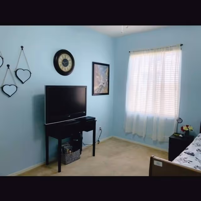 A bedroom with light blue walls, a window with white curtains letting in natural light, a black TV on a small black table, a wall clock above the TV, two heart-shaped mirrors hanging on the wall, a framed picture on the wall, and a bed partially visible with floral bedding and a nightstand with a lamp and flowers.