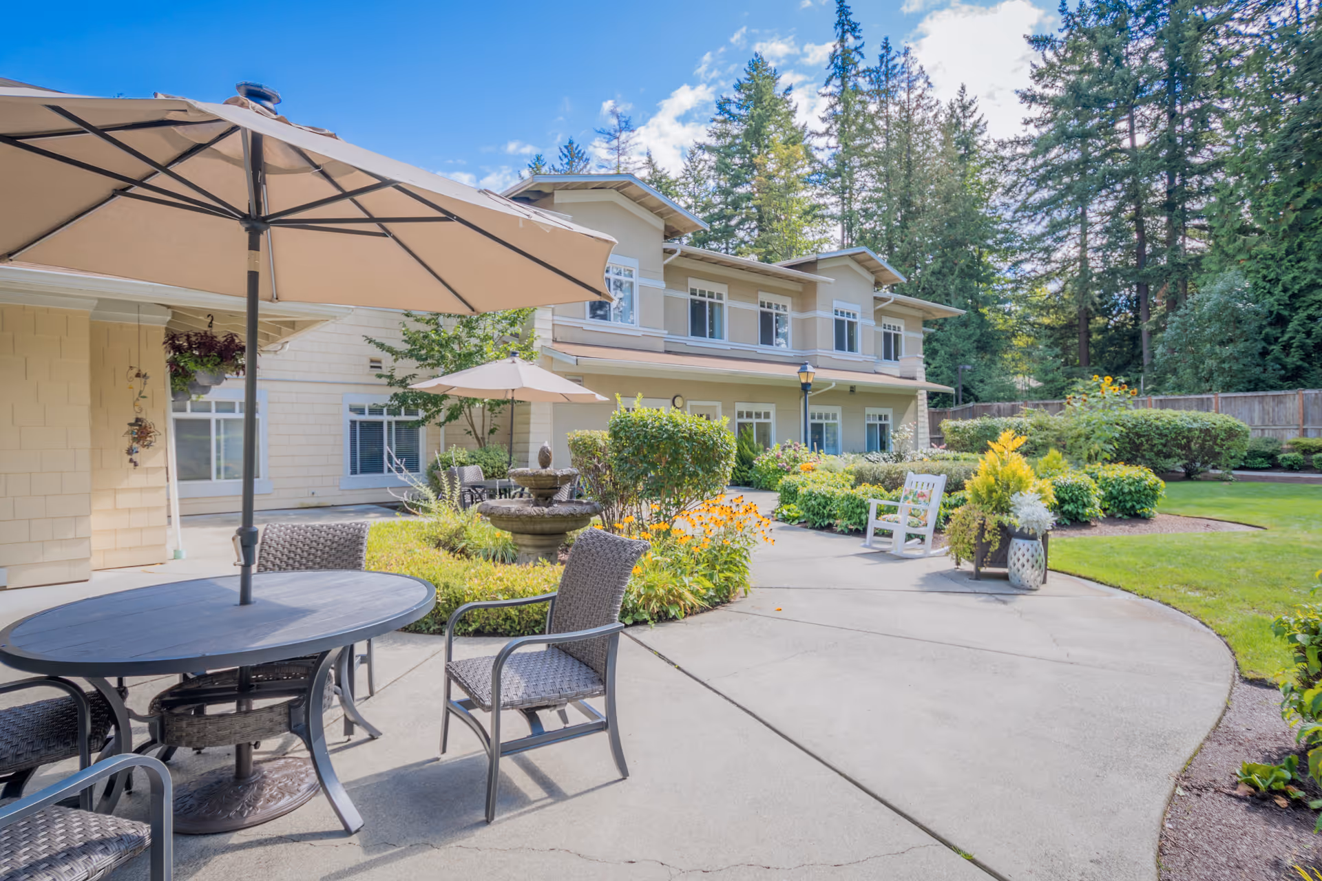 Outdoor patio area at Sunrise of Redmond with round tables and chairs under large beige umbrellas, surrounded by landscaped greenery, flowers, and a two-story building in the background under a blue sky with some clouds.