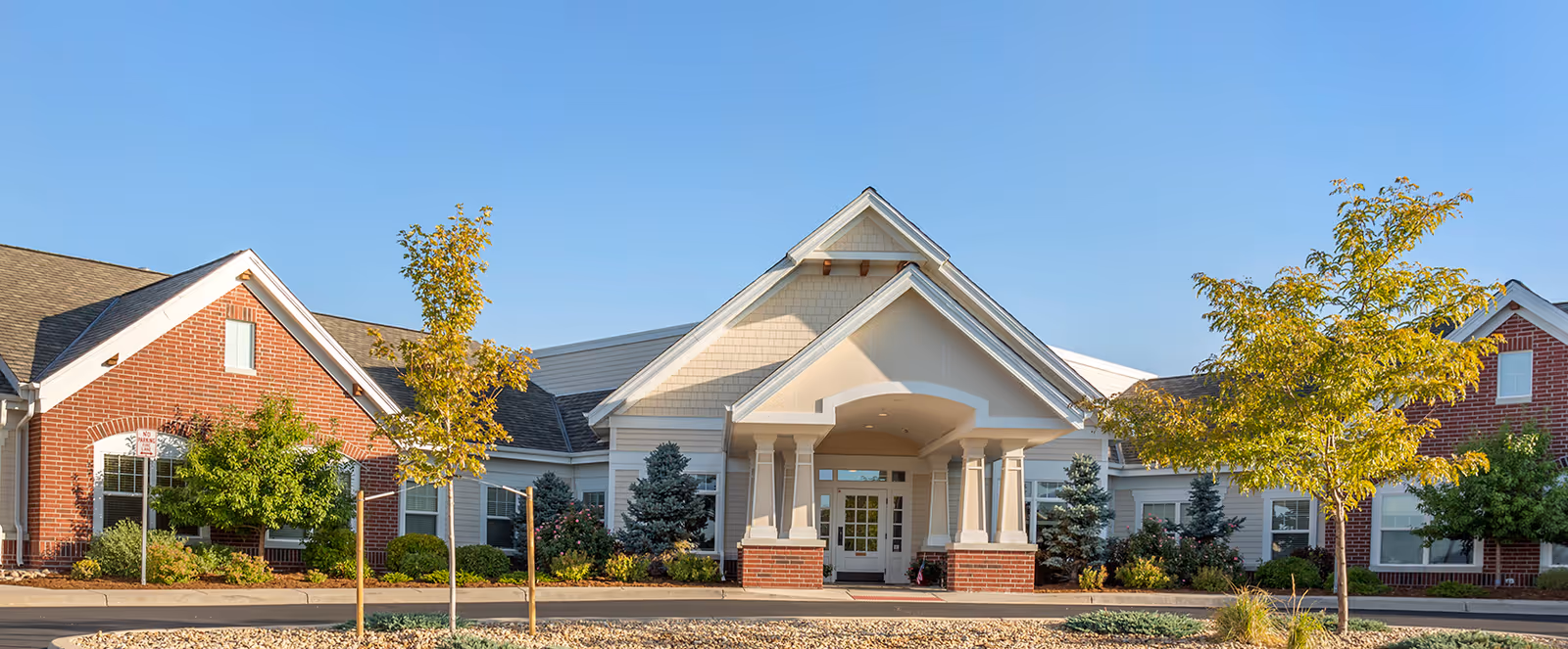 Front exterior view of Brookdale Highlands Ranch facility showing a single-story building with a combination of brick and light-colored siding, a covered entrance with columns, and landscaped greenery including trees and shrubs under a clear blue sky.