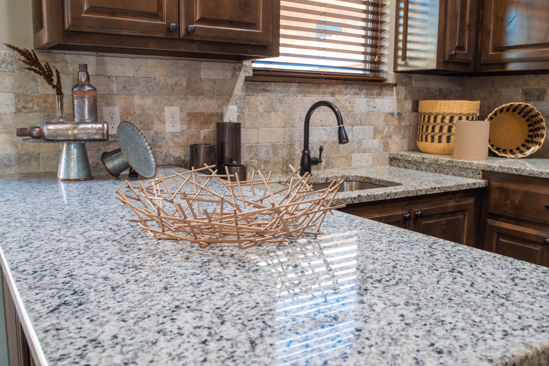 A kitchen countertop with a decorative twig basket in the center. The countertop is granite with a speckled pattern. Behind the counter are dark wooden cabinets, a black faucet, a small sink, and a window with wooden blinds. Various decorative items, including a metal sculpture and woven baskets, are placed on the counter and against the tiled backsplash.