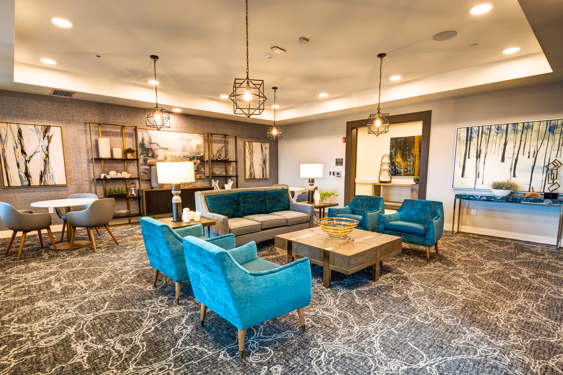 Spacious, well-lit senior living lounge with teal armchairs, a gray sofa, coffee table, artwork and pendant lights.