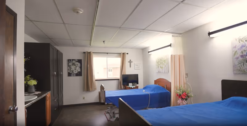 A simple, clean room with two hospital-style beds covered in blue blankets. The room has a window with beige curtains, a small TV on a stand, a cross on the wall, and floral artwork. There is a cabinet with a sink on the left side and a bouquet of flowers near one of the beds.