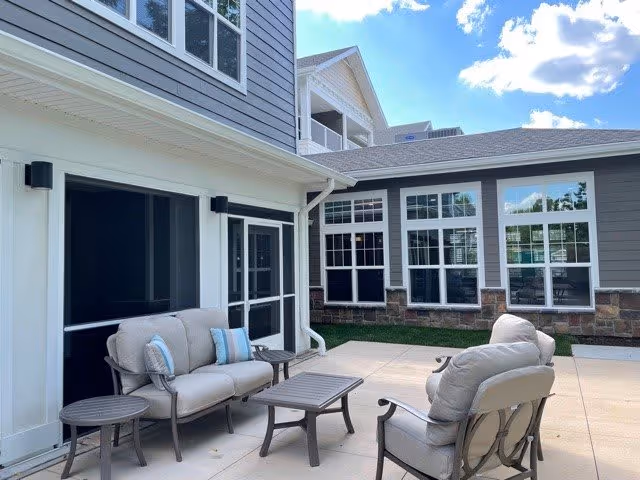 Sunny outdoor courtyard with cushioned patio chairs and tables in front of a gray-sided senior living building.