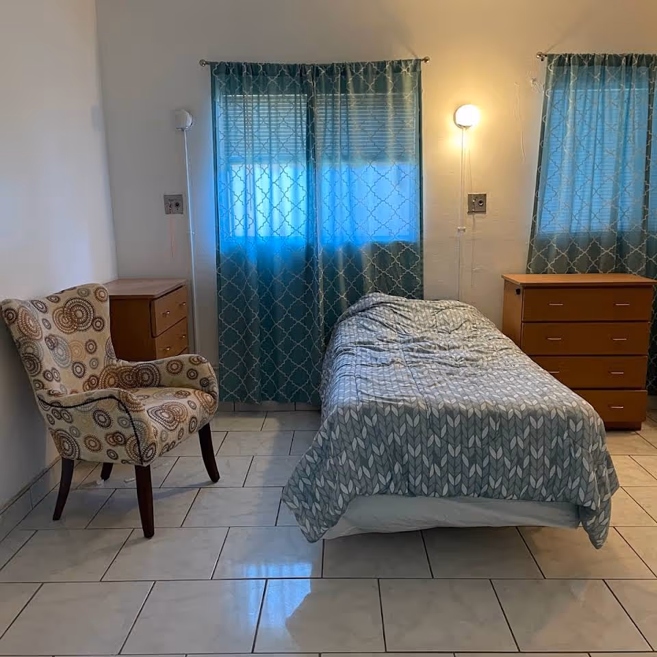 A simple bedroom with a single bed covered in a gray patterned comforter, a wooden dresser on the right, a small wooden nightstand on the left, a patterned upholstered armchair, and two windows with blue patterned curtains letting in soft light.