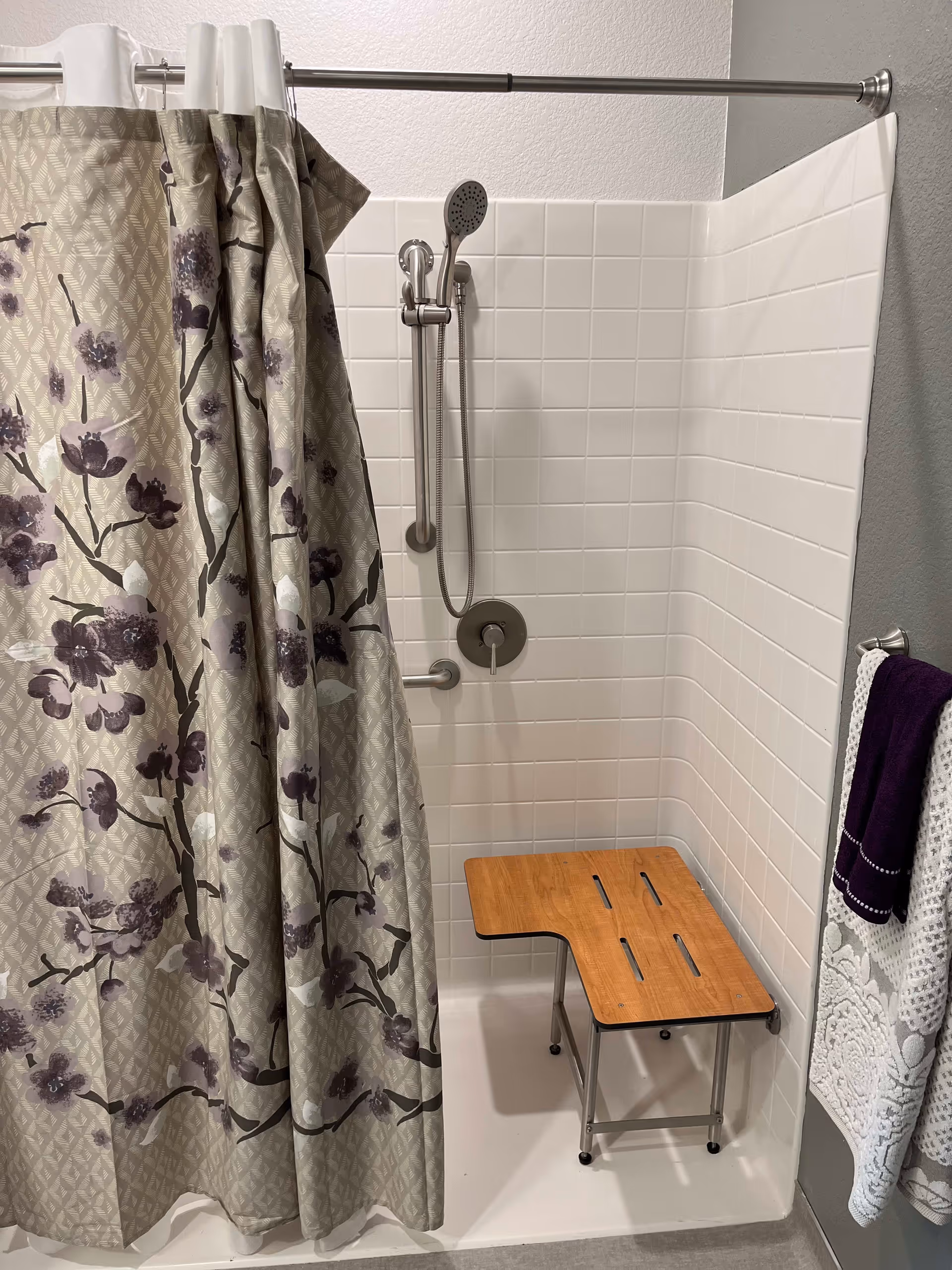 Shower stall with a floral curtain, handheld showerhead, grab bar, and a wooden shower bench.