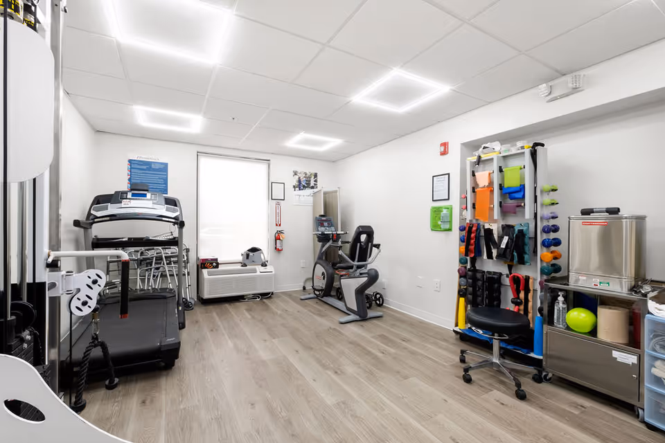 Small fitness/therapy room with a treadmill, stationary bike, weight and resistance equipment, and storage carts.