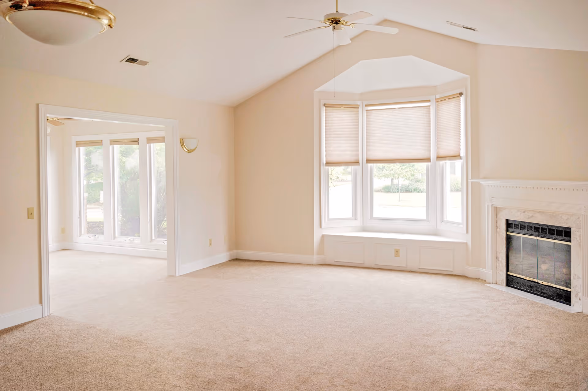 A bright and empty living room with beige carpet, a white ceiling fan, a bay window with closed blinds, and a fireplace with a marble surround. Adjacent to the living room is another room with large windows letting in natural light.