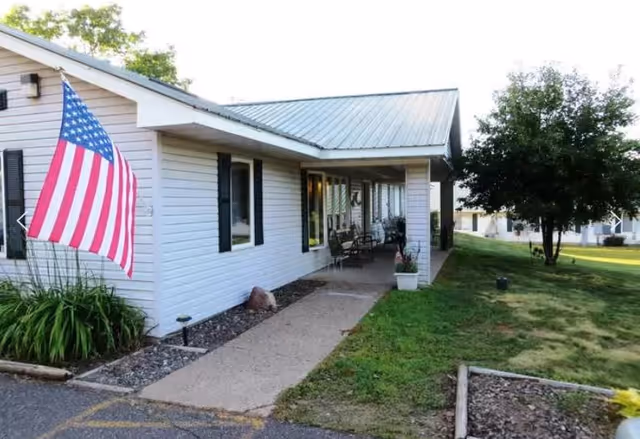 Exterior view of a single-story building with white siding and a metal roof. An American flag is mounted near the corner of the building. There is a paved walkway leading to a covered porch area with chairs and potted plants. The surrounding area includes grass, trees, and landscaping with rocks.