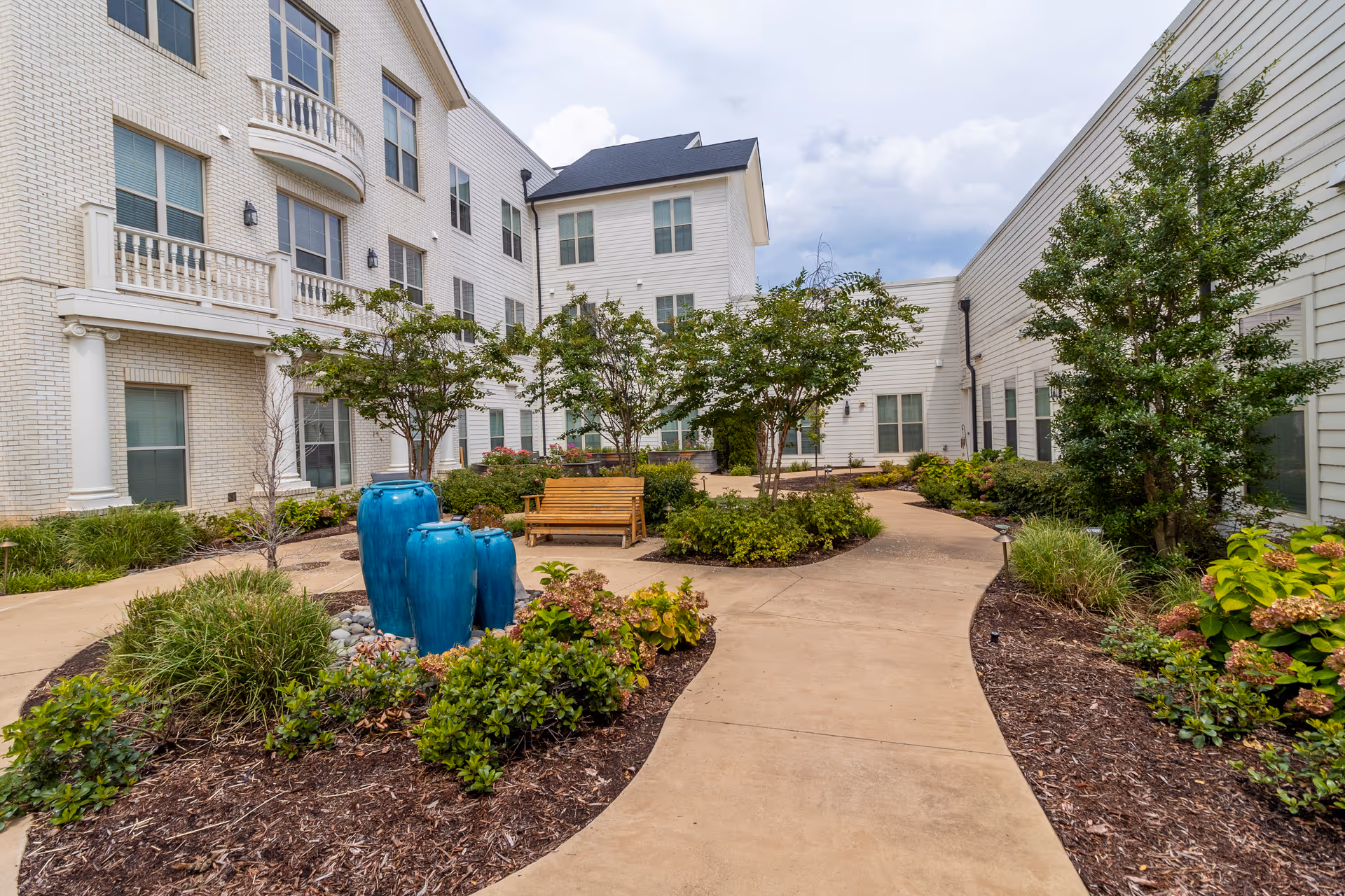 Outdoor courtyard area at StoryPoint Germantown featuring a winding concrete pathway, landscaped garden beds with shrubs and small trees, three large blue ceramic water fountains, and a wooden bench. The courtyard is surrounded by a white multi-story building with multiple windows.