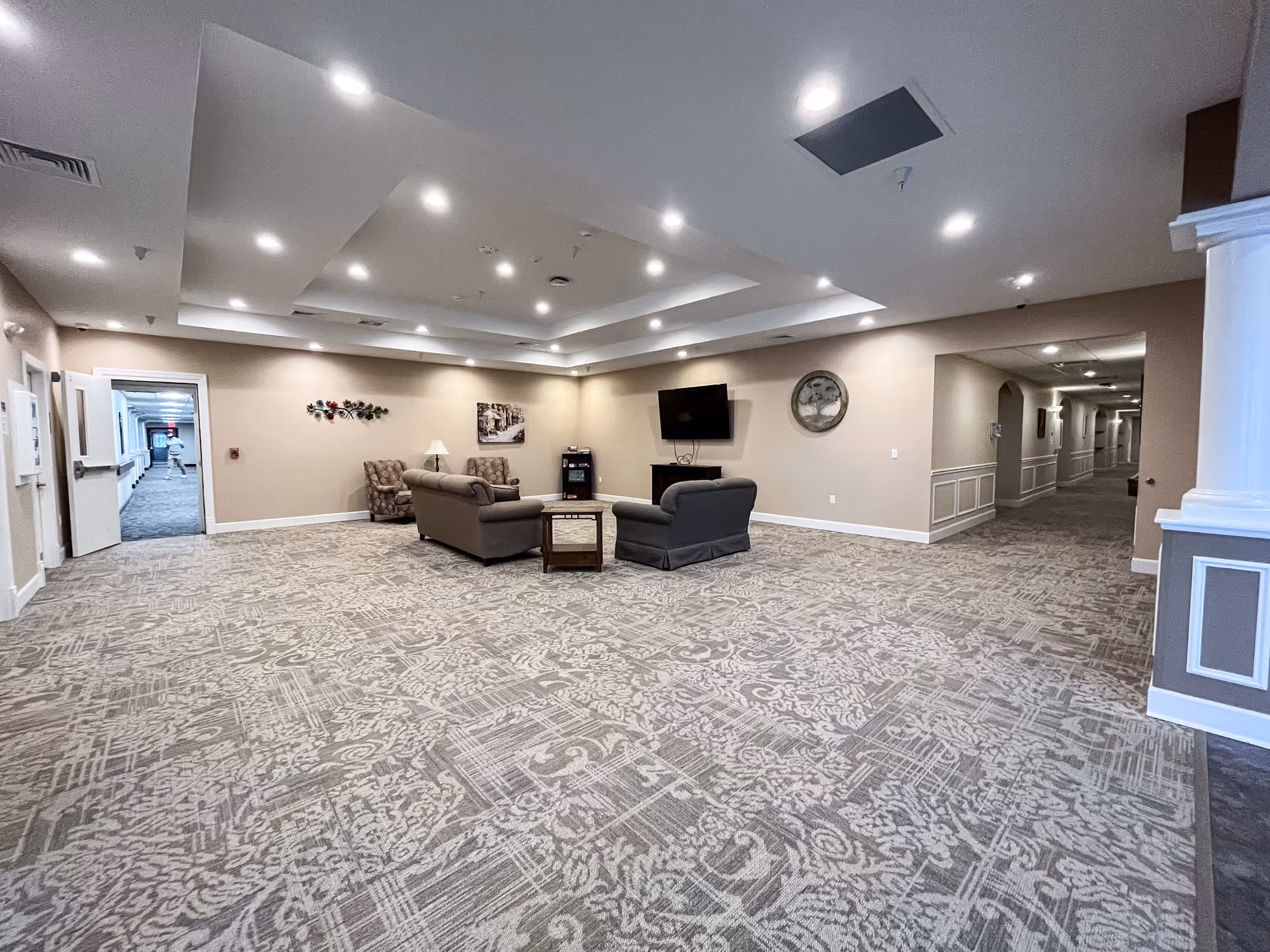 A spacious common area in a senior living facility with patterned carpet flooring, beige walls, and recessed ceiling lights. The room features a seating arrangement with two sofas, two armchairs, a coffee table, a side table with a lamp, a wall-mounted TV, and decorative wall art. Two hallways extend from the room, one with an open door showing a corridor with a person walking.