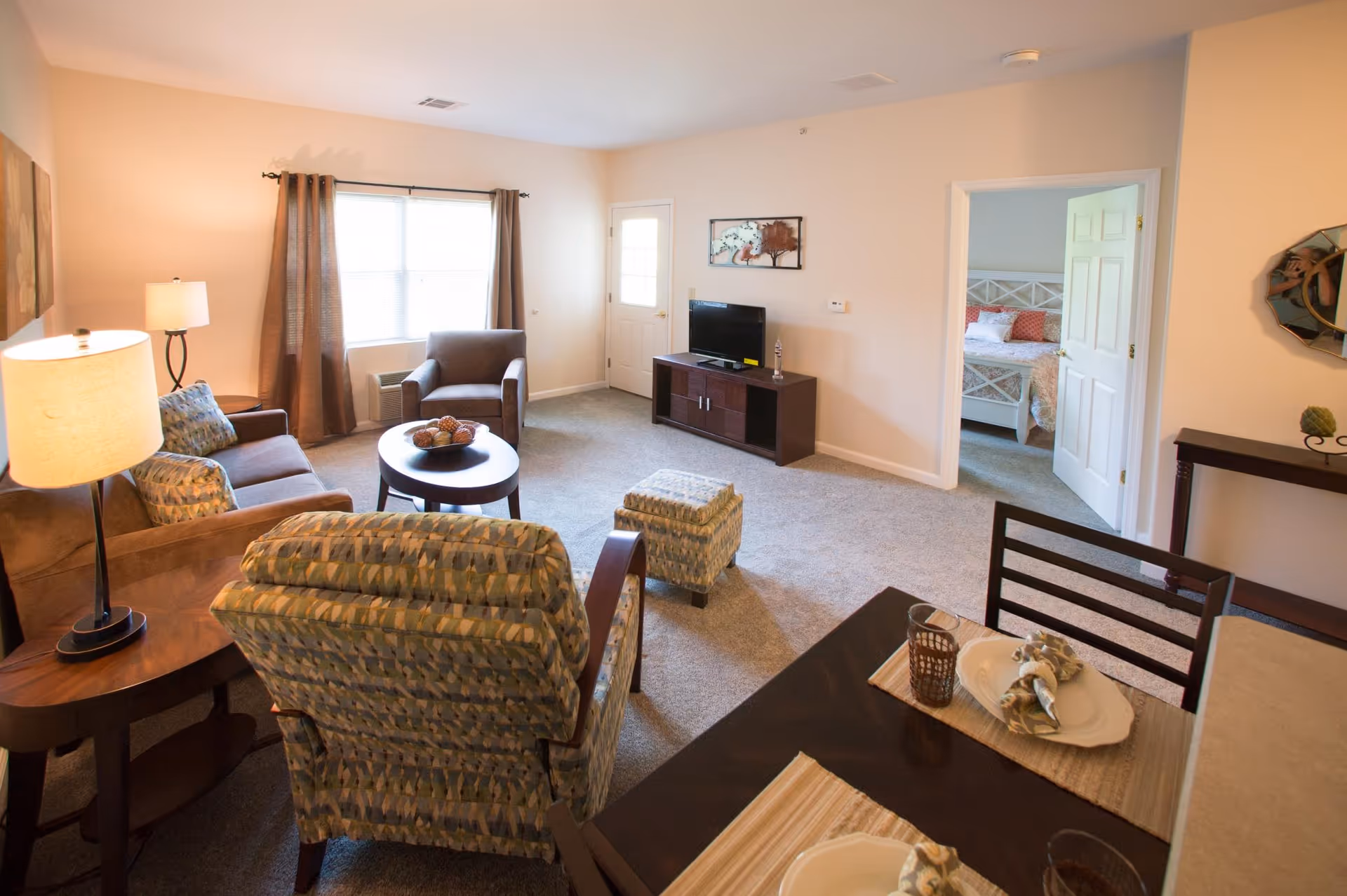 A bright living room with beige walls and carpeted floor. The room features a patterned armchair with matching ottoman, a brown sofa with patterned cushions, a round coffee table with decorative items, a side table with a lamp, and a TV on a dark wooden stand. A window with brown curtains lets in natural light. A doorway leads to a bedroom with a white bed frame and colorful bedding. A dining table with place settings is partially visible in the foreground.