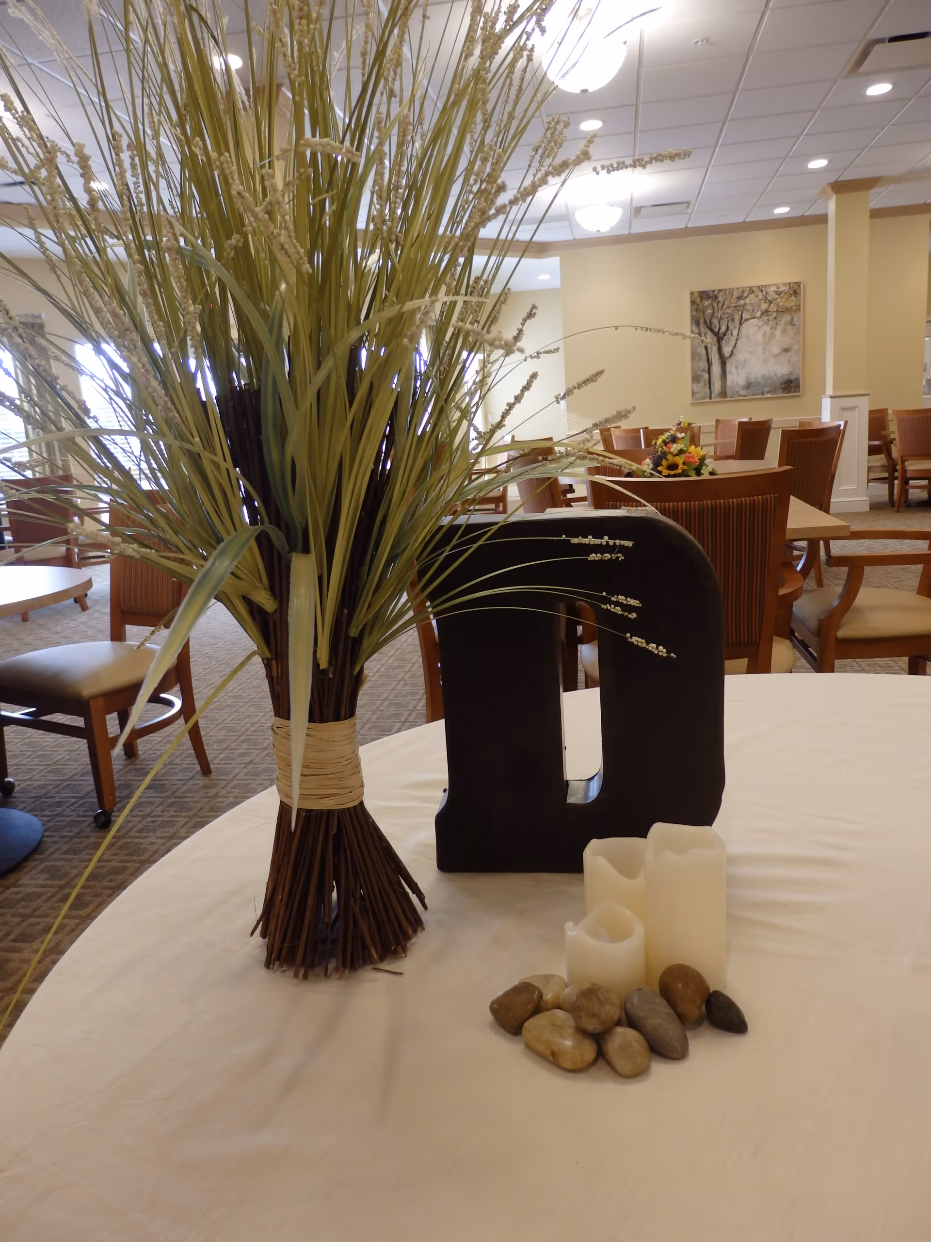 A round table with a white tablecloth displaying a decorative arrangement including a bundle of tall grass tied with twine, a large black letter 'D', three white candles, and several small stones. In the background, there are wooden chairs and tables in a well-lit dining area with beige walls and a framed painting.