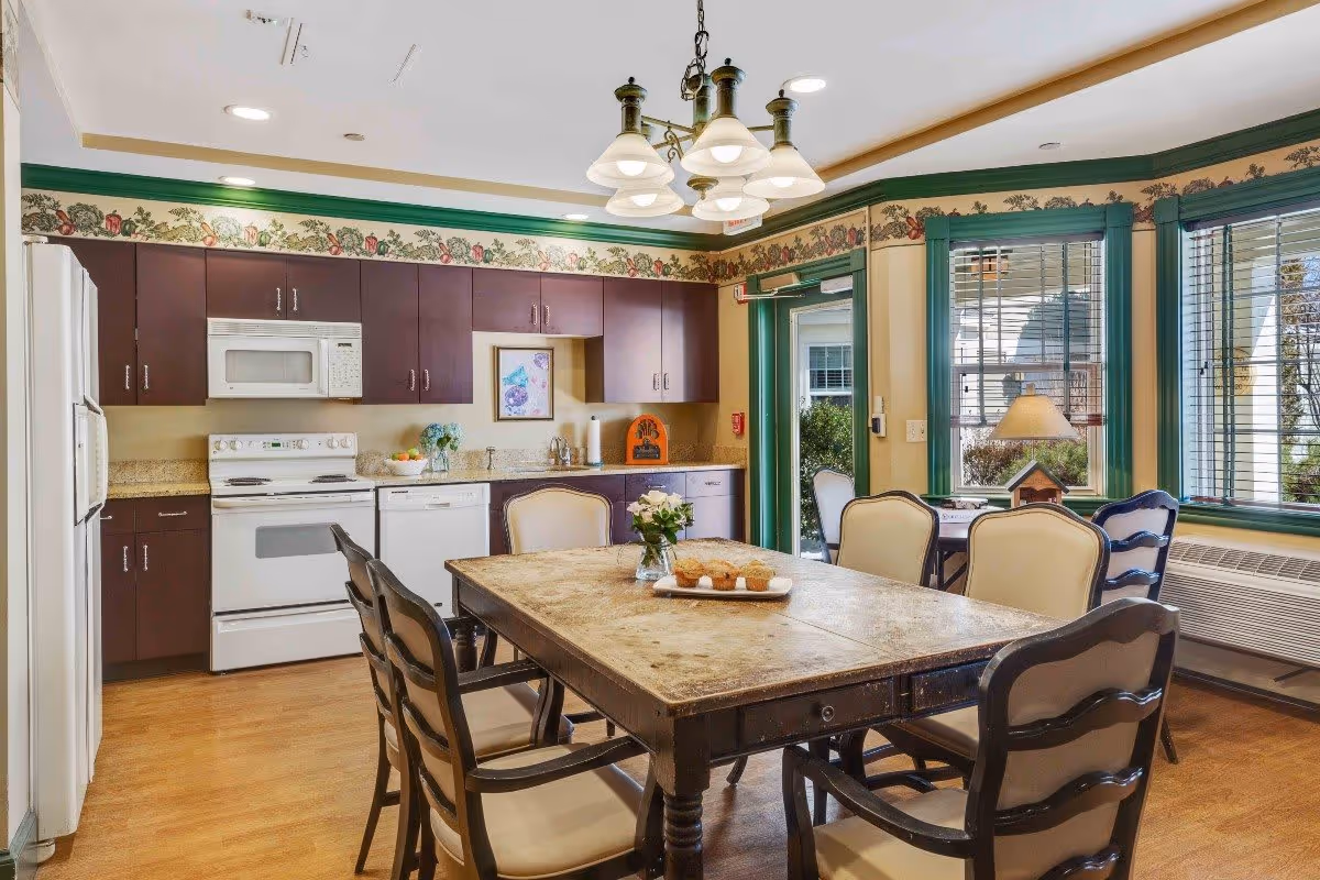 A bright kitchen and dining area in a senior living facility featuring wooden cabinets, white appliances including a refrigerator, stove, microwave, and dishwasher. A wooden dining table with six cushioned chairs is centered in the room, with a small vase of flowers and a tray of muffins on the table. The room has large windows with green trim, a hanging light fixture above the table, and floral wallpaper border near the ceiling.